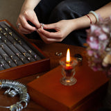 Person handling jewelry near a wooden box with rings and a lit candle on a table.
