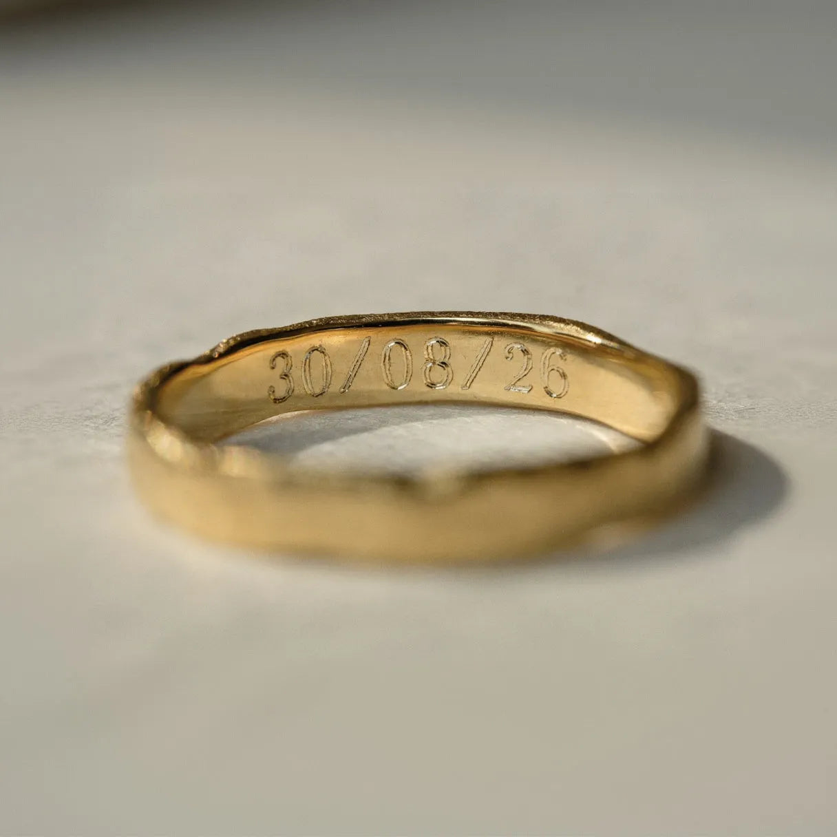 A close-up of a gold band ring with a textured, organic shape and an engraved date '30/08/26' on the inside, resting on a light gray surface.