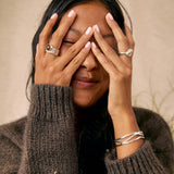 A woman with her hands over her eyes wearing a dark brown ribbed knit sweater, a black turtleneck, and silver rings and bracelets, set against a warm beige textured background.