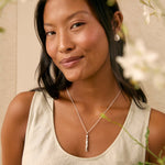 A woman with dark hair wearing a silver bar pendant necklace and a light beige tank top, posing with soft-focus white flowers in the foreground.