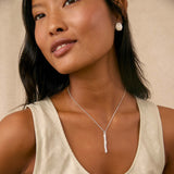 A woman wearing a chunky silver necklace with a vertical bar pendant and a spiral silver stud earring, paired with a light beige linen tank top against a neutral beige background.