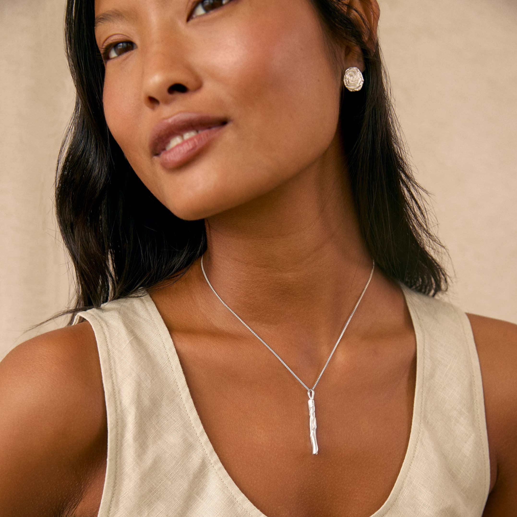 A woman wearing a chunky silver necklace with a vertical bar pendant and a spiral silver stud earring, paired with a light beige linen tank top against a neutral beige background.