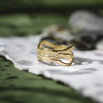 A gold ring with a wavy, organic, tree-bark-like design, resting on a white textured piece of paper against a dark green, softly blurred background.