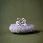 A silver ring with a twisted, organic shape resting on a smooth, light purple stone, set against a dark olive green background.