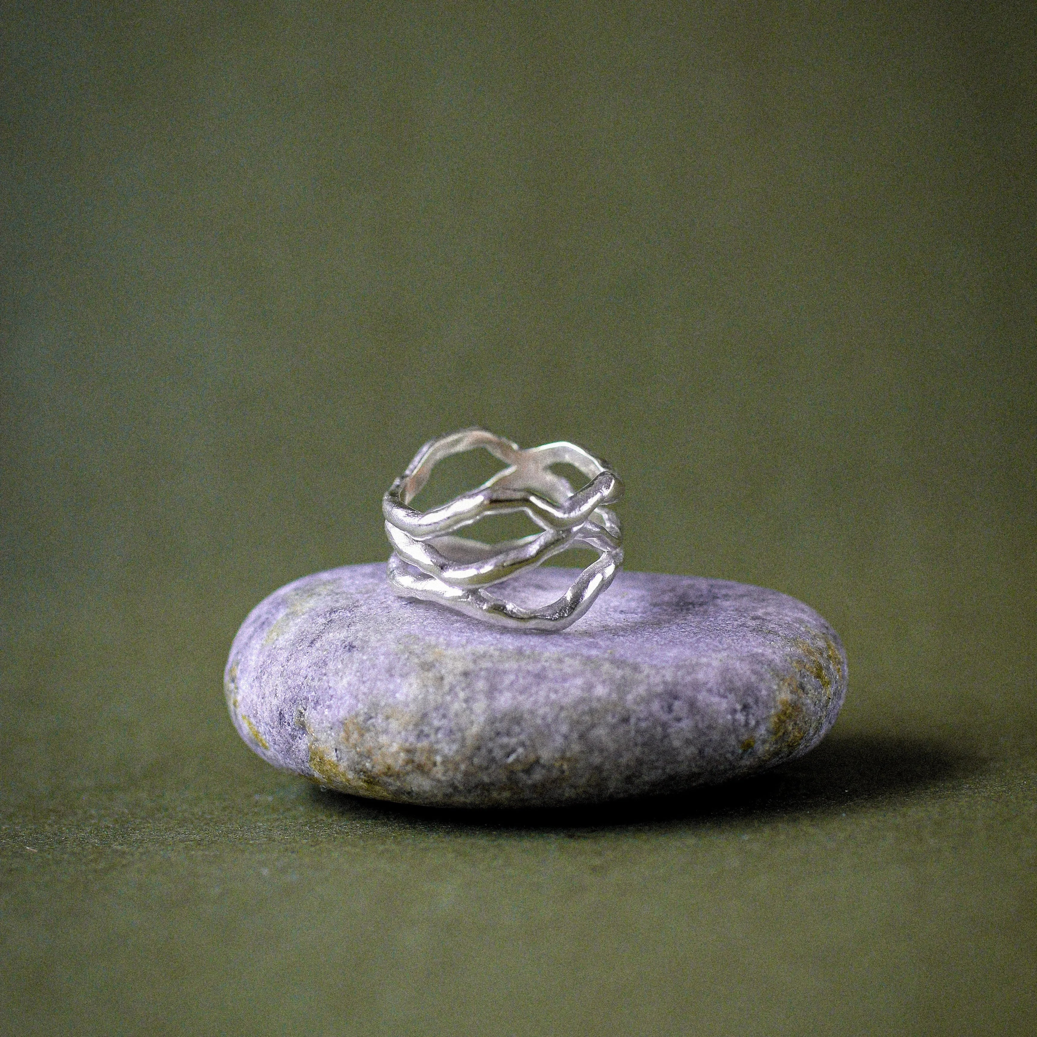 A silver ring with a twisted, organic shape resting on a smooth, light purple stone, set against a dark olive green background.