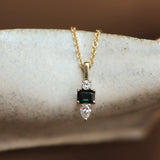a delicate gold necklace with square green sapphire and round and pear diamonds hanging on the edge of a ceramic dish