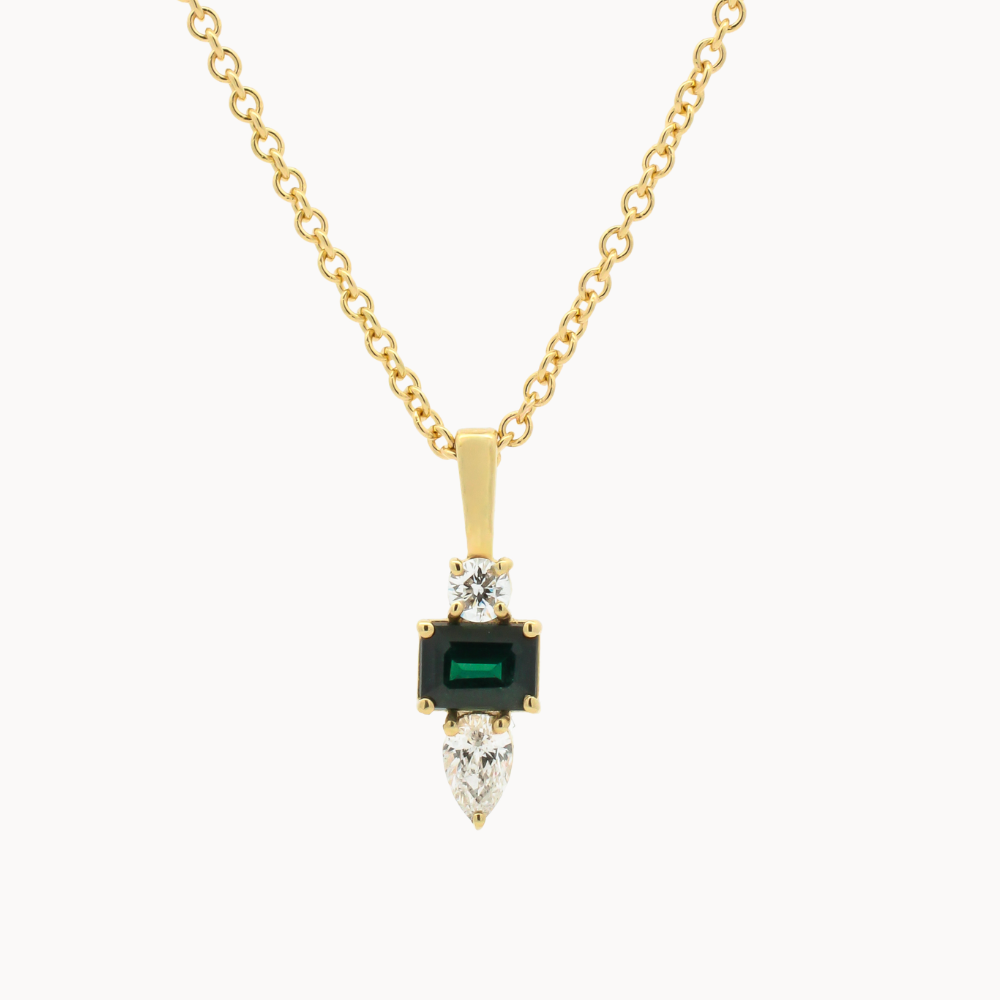 A gold pendant necklace featuring a central rectangular green emerald-cut stone, flanked by clear marquise and round stones, suspended from a delicate gold chain on a light gray background.