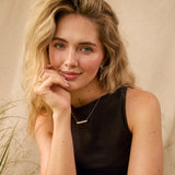 A blonde woman posing in a black sleeveless top and beige skirt, wearing a delicate gold bar necklace, set against a warm beige backdrop with dried grasses.