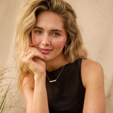 A blonde woman posing in a black sleeveless top and beige skirt, wearing a delicate silver bar necklace, set against a warm beige backdrop with dried grasses.