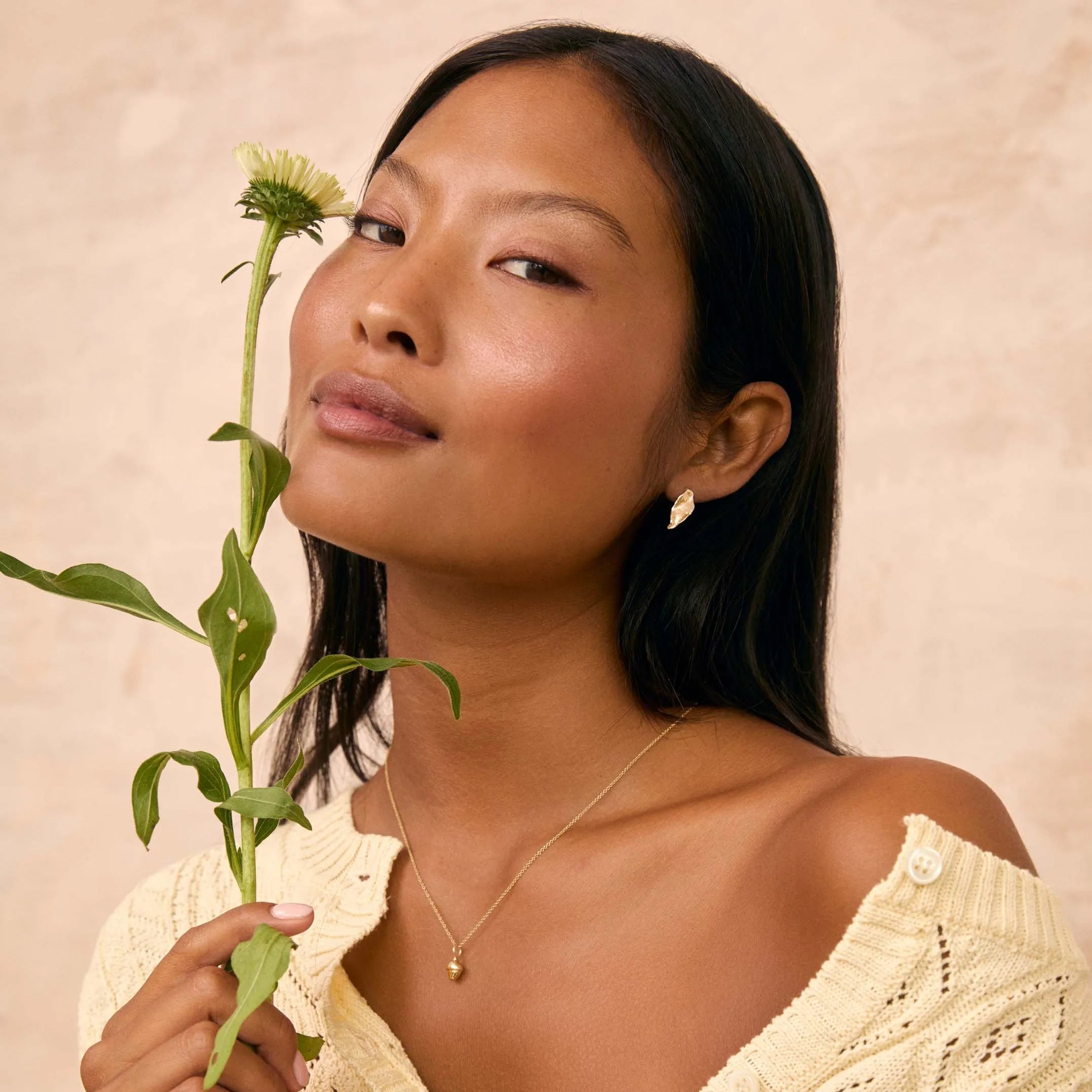 A woman with dark hair wearing a cream off-the-shoulder knit sweater and a delicate gold acorn necklace and leaf earrings, holding a green flower stem against a textured beige background.