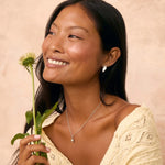 A woman with long black hair smiles while looking upward, holding a small green flower. She wears a cream knit top, a delicate silver acorn necklace and leaf earrings against a warm beige textured background.