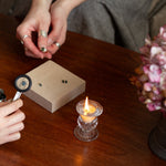 Two people examining gemstones and engagement rings with a magnifying glass on a wooden table, candle and water glass in the background.