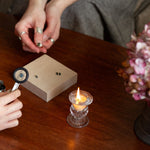 Two people examining gemstones and engagement rings with a magnifying glass on a wooden table, candle and water glass in the background.