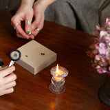 Two people examining gemstones and engagement rings with a magnifying glass on a wooden table, candle and water glass in the background.