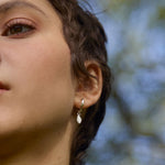 Close-up of a person wearing a gold oak leaf earring with a blurred natural background of trees