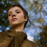 Woman with short hair wearing a brown cargigan and oak leaf earrings against a blurred leafy background