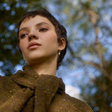 Woman with short hair wearing a brown cargigan and oak leaf earrings against a blurred leafy background