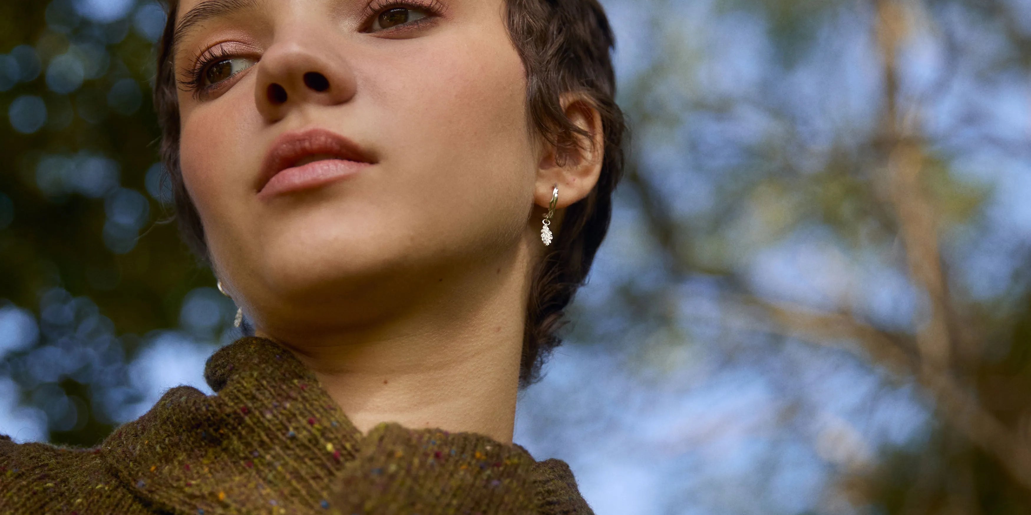 Woman with short hair wearing a brown cargigan and oak leaf earrings against a blurred leafy background