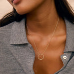 A close-up of a model wearing a delicate gold necklace with a minimalist circular pendant, styled with a gray button-up shirt.