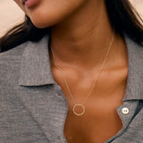 A close-up of a model wearing a delicate gold necklace with a minimalist circular pendant, styled with a gray button-up shirt.