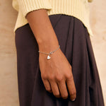A close-up of a person wearing a delicate silver chain bracelet with a small heart-shaped charm, styled with a cream knit sweater and a dark brown skirt against a textured beige wall.