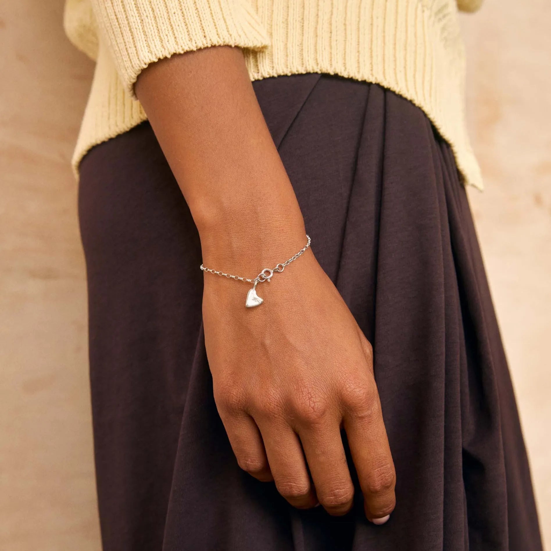 A close-up of a person wearing a delicate silver chain bracelet with a small heart-shaped charm, styled with a cream knit sweater and a dark brown skirt against a textured beige wall.