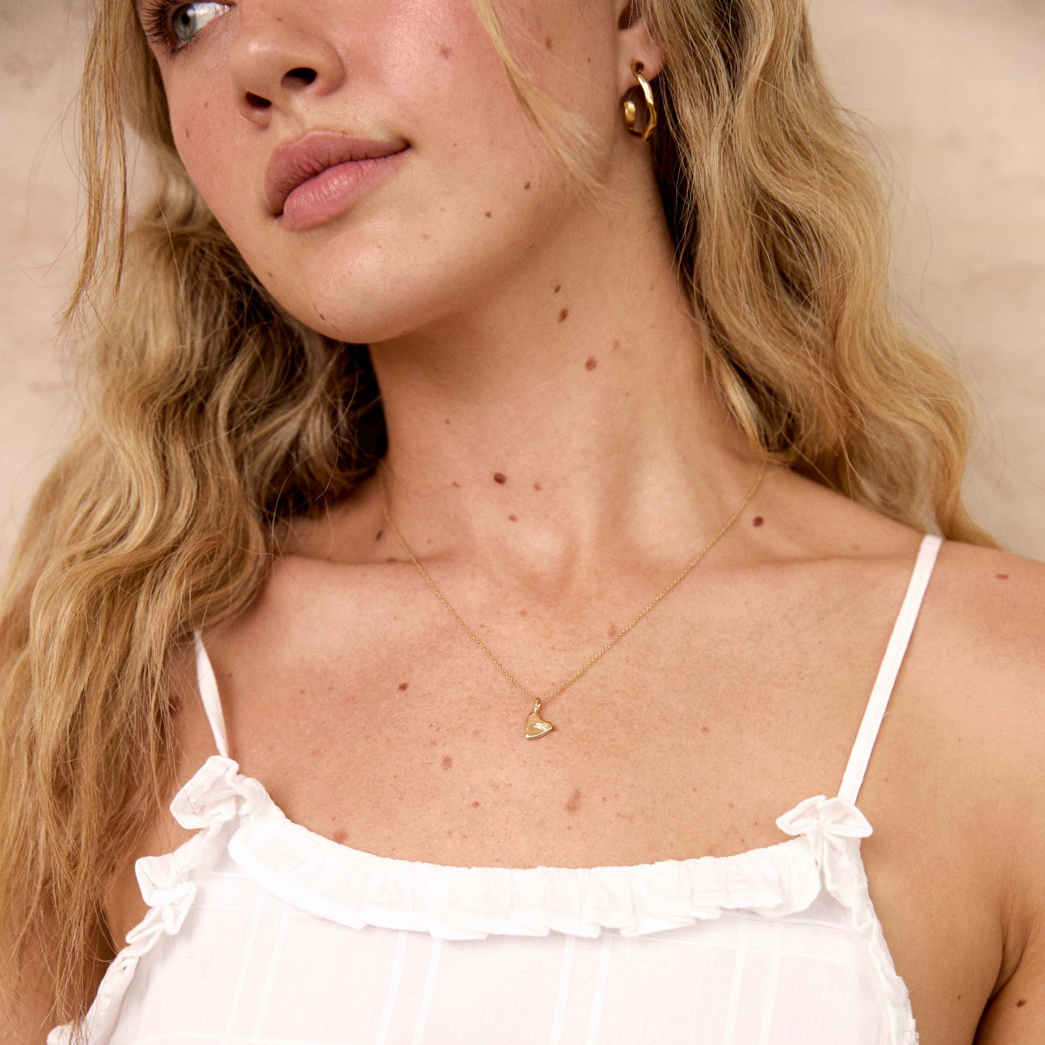 A close-up of a blonde woman wearing a delicate silver necklace with a small heart pendant, paired with a white ruffled spaghetti strap top.