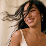 Smiling woman with flowing hair wearing a strappy wedding dress, gold earrings and necklace against a neutral background