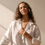 Woman in a white shirt and silver heart necklace sitting on the floor with sunlight casting shadows.