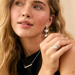 A blonde woman wearing a black sleeveless top and silver jewelry, including a ring and necklace, posing in front of a neutral beige backdrop with green plants.