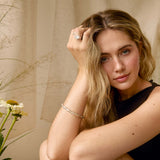 A woman with blonde hair sits cross-legged in a warm, neutral-toned setting, wearing a black sleeveless top, a silver bracelet, and a ring. She is framed by dried grasses and white daisies on both sides.