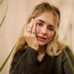 A woman with blonde hair wearing a dark brown ribbed knit sweater and silver rings, posing with dried grass plants in the foreground.