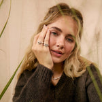 A woman with blonde hair wearing a dark brown ribbed knit sweater and silver rings, posing with dried grass plants in the foreground.