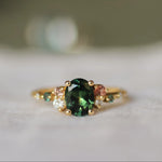engagement ring with large green sapphire and smaller pink, teal sapphires and round diamonds with a blurred background