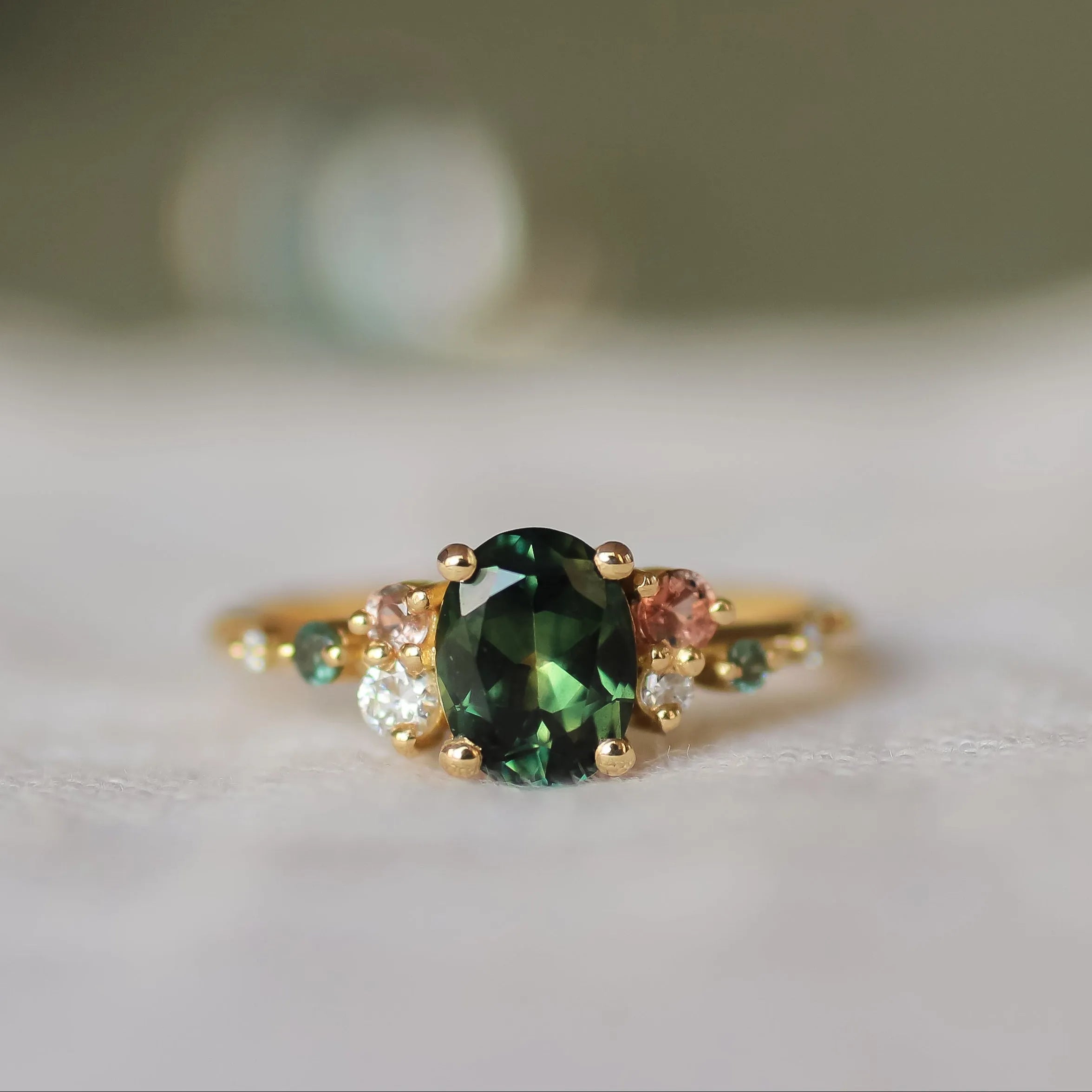 engagement ring with large green sapphire and smaller pink, teal sapphires and round diamonds with a blurred background