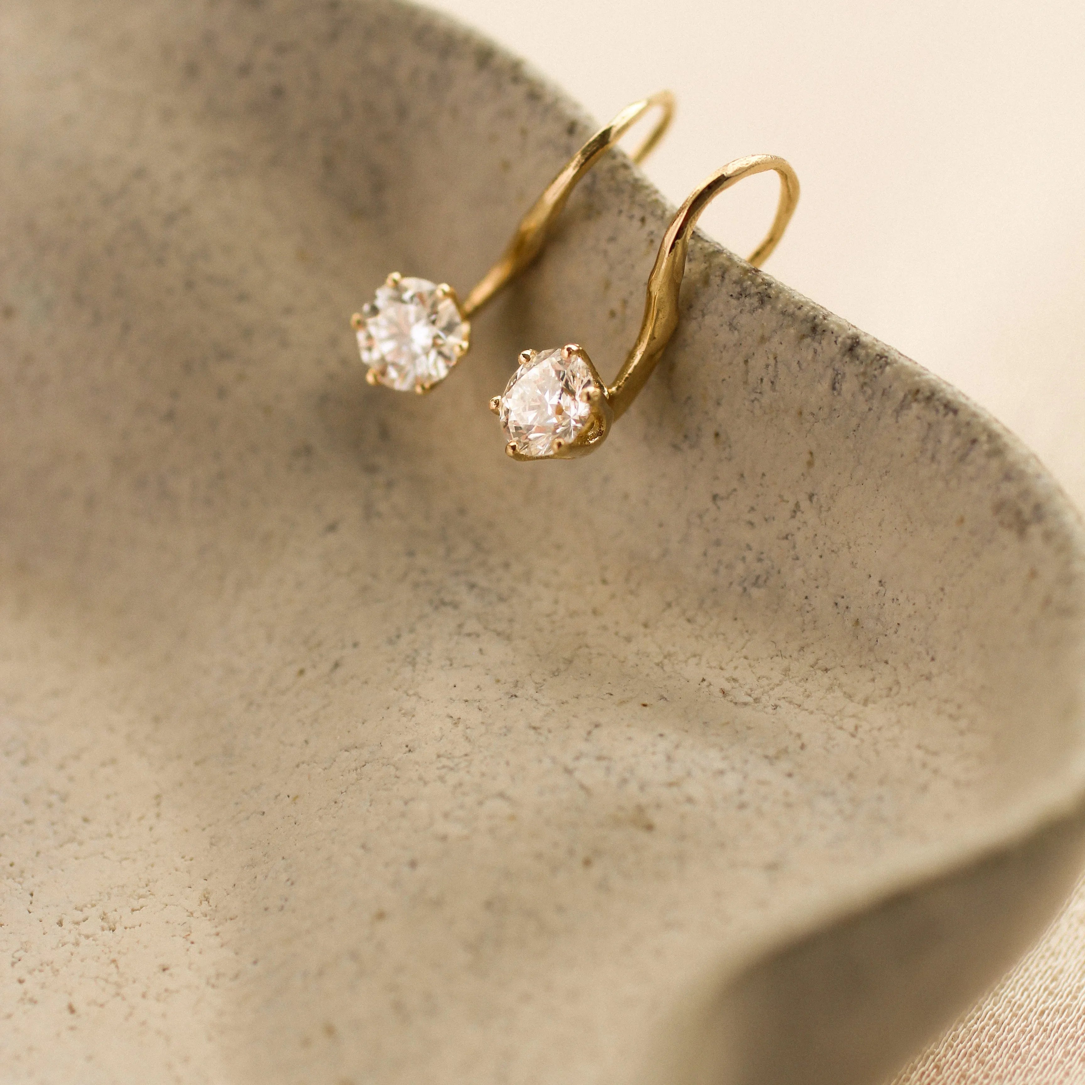 Gold earrings with round diamonds hooked onto the edge of a beige ceramic dish