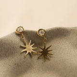 Gold sun-shaped earrings with clear gemstones on a beige ceramic surface