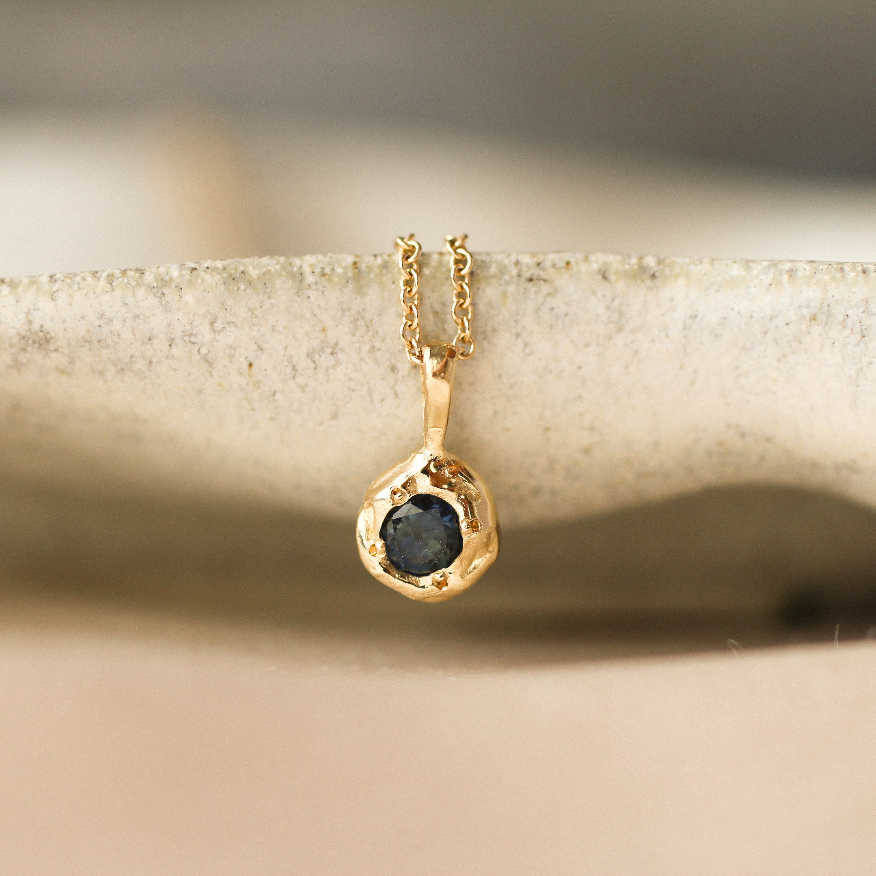 Gold necklace with a pendant featuring a dark blue gemstone hanging from the edge of a beige ceramic dish