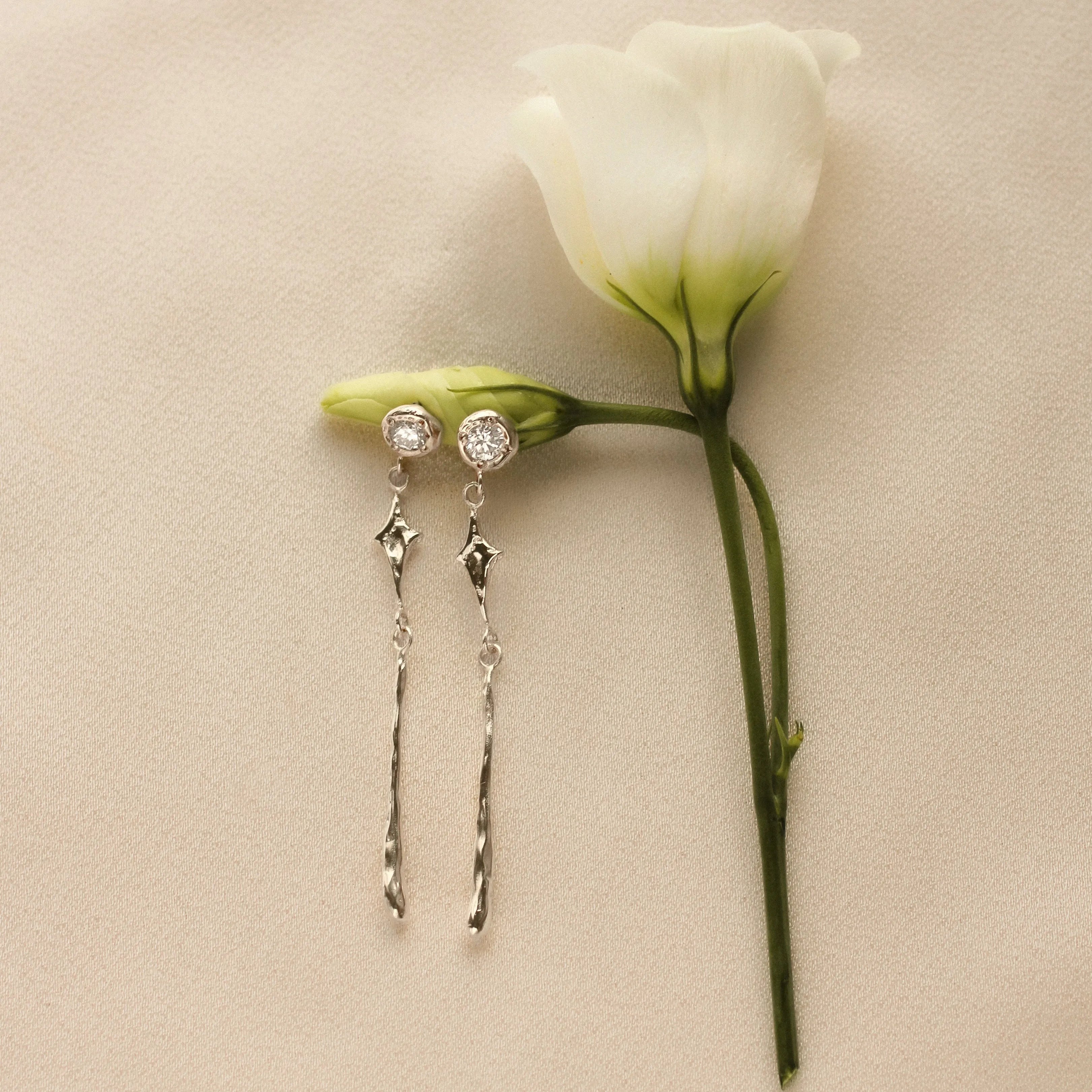 Pair of silver-toned gold earrings with diamond, star and molten detailing on a white rose step with a beige background