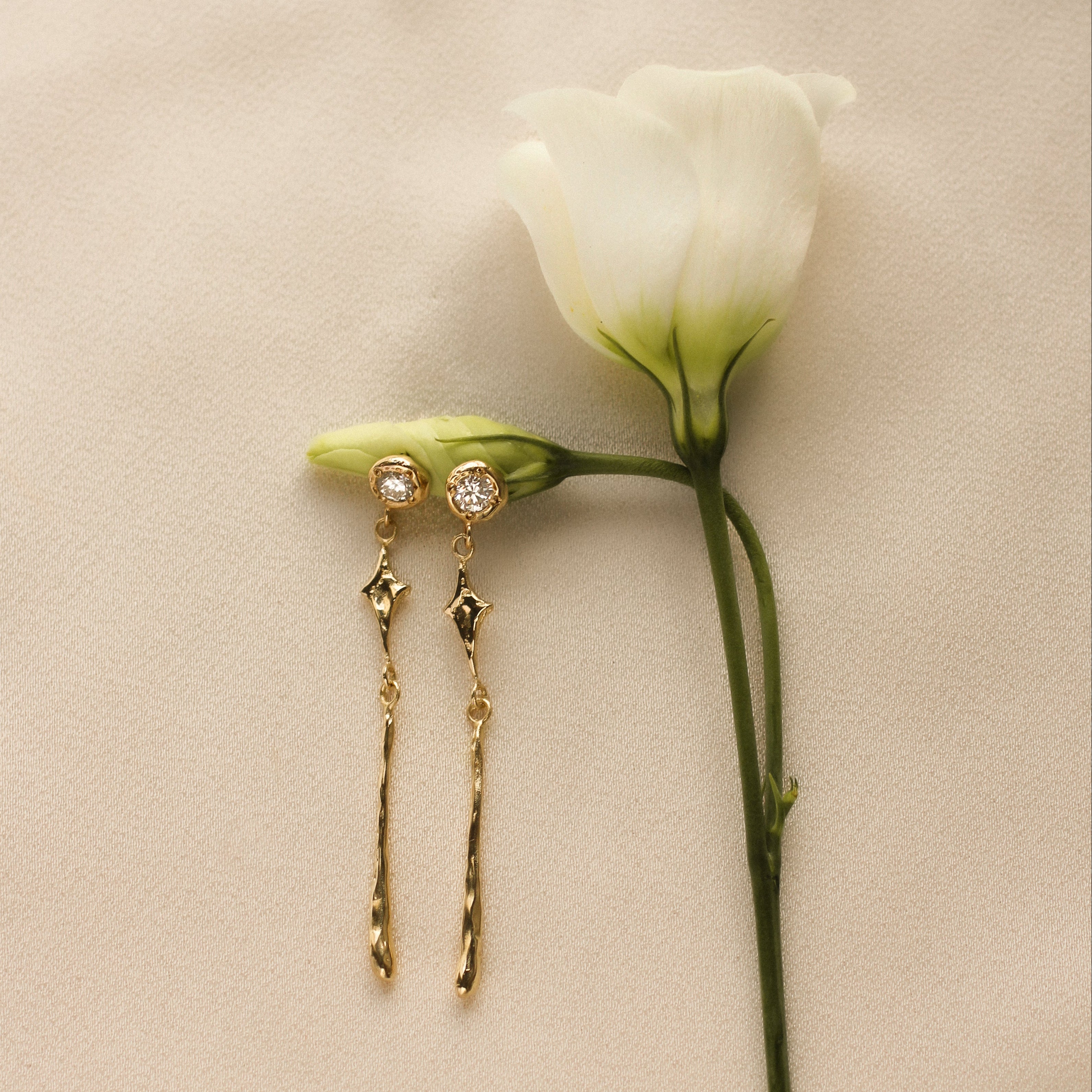 Gold earrings with diamond and star details next to a white flower on a beige background