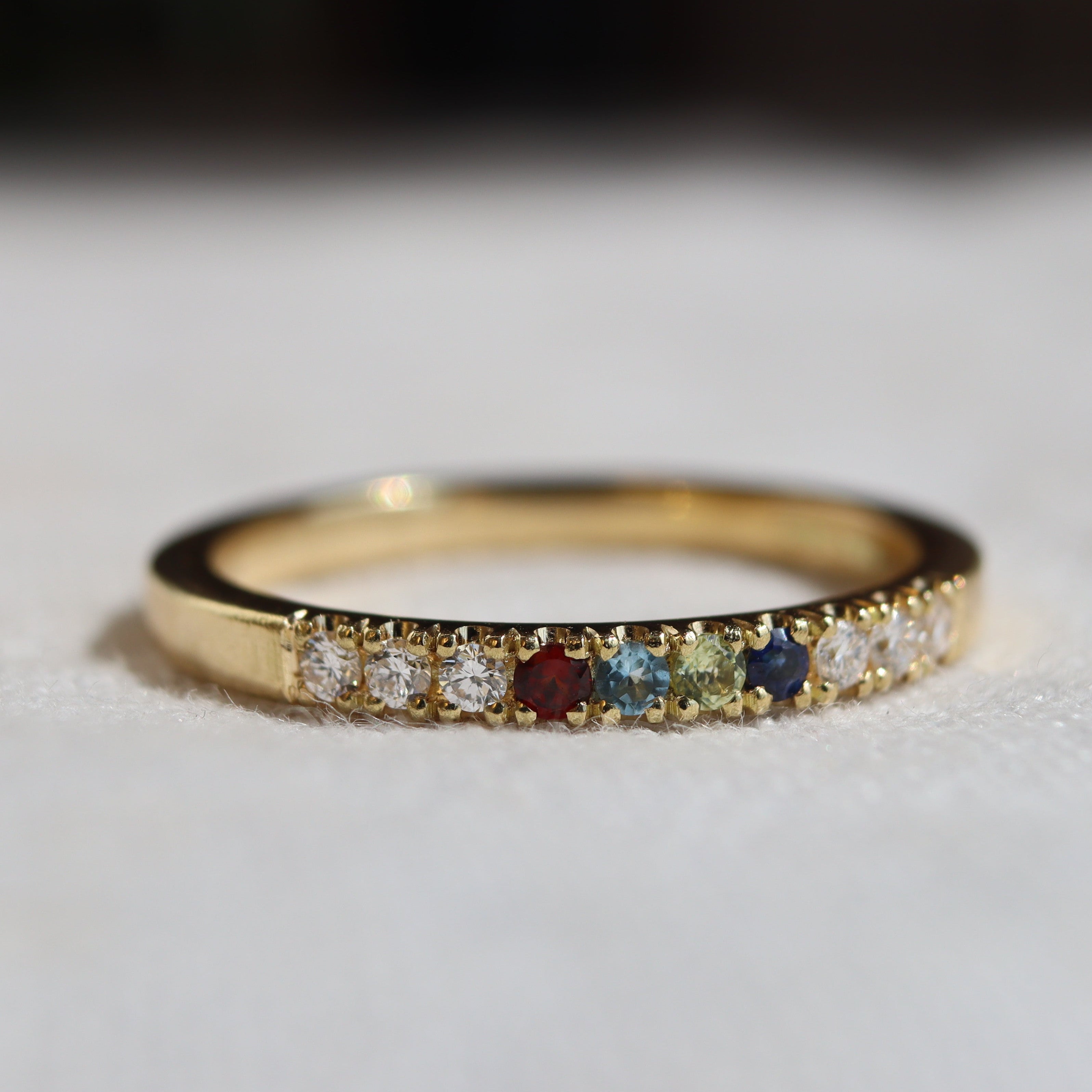 A yellow gold eternity ring featuring a row of small multicolored gemstones, including clear, red, blue, and green stones, resting on a textured white fabric surface.