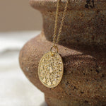A gold round pendant necklace with an intricate moonphase design hangs from a gold chain, draped over the rim of a rustic brown ceramic pot on a light beige fabric surface.