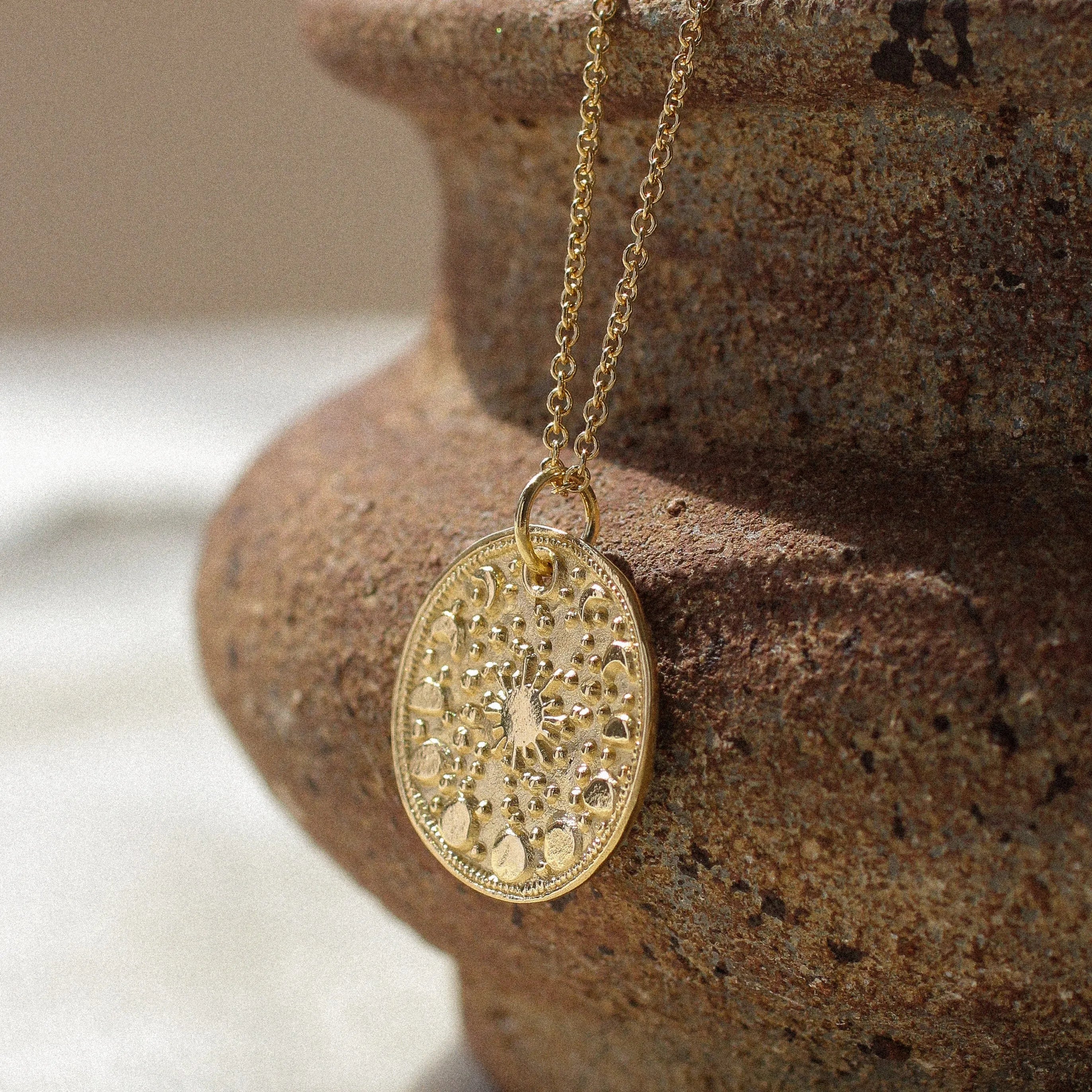 A gold round pendant necklace with an intricate moonphase design hangs from a gold chain, draped over the rim of a rustic brown ceramic pot on a light beige fabric surface.