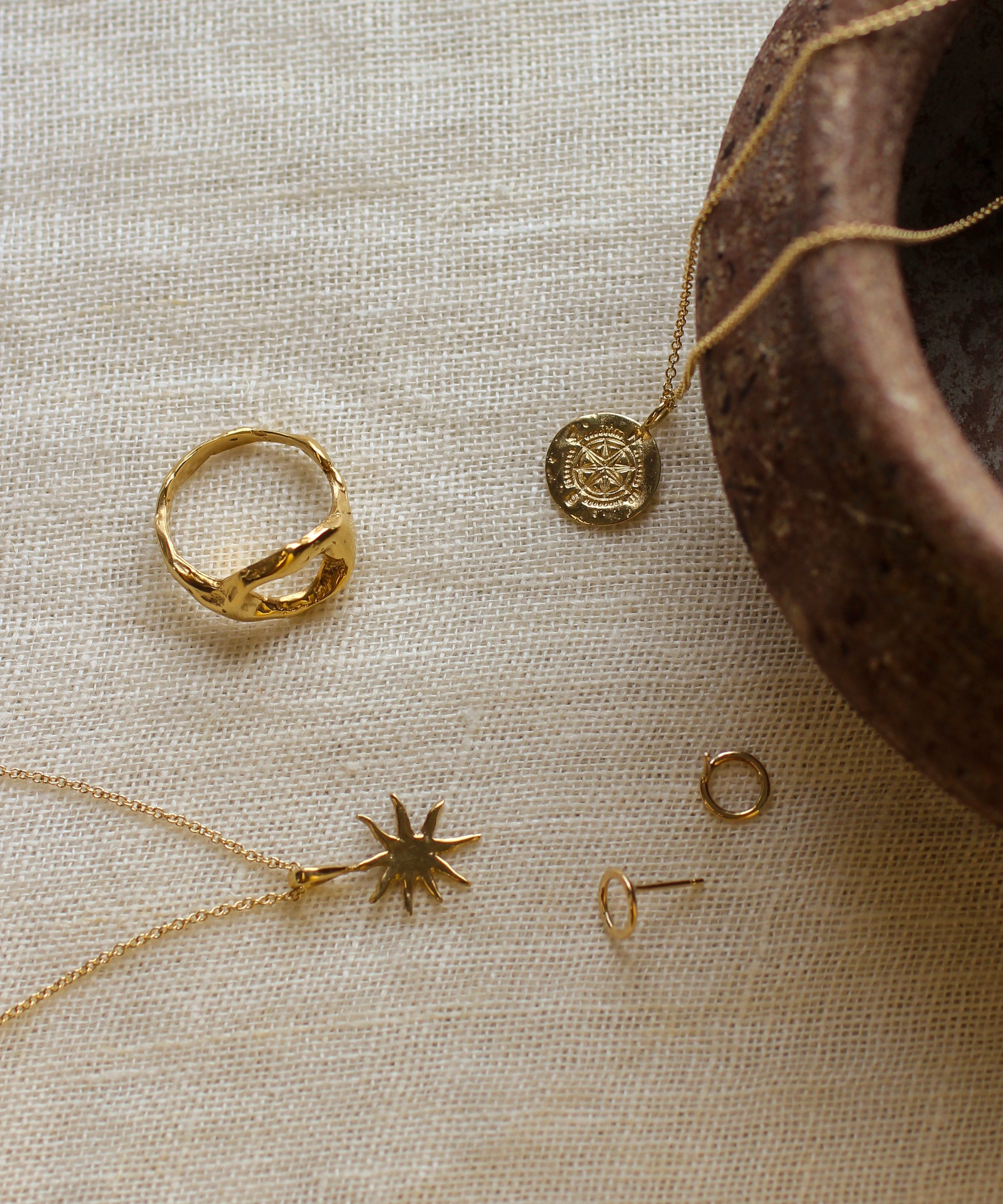 Gold jewelry including a ring, necklace, and earrings on a textured beige surface with a brown ceramic bowl.