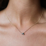 A close-up of a delicate gold chain necklace worn by a model, featuring a greeny blue sapphire and pear shaped diamond.