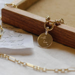 Gold necklace pendant on a chunky chain with a 'with love' charm on a wooden box with a white background