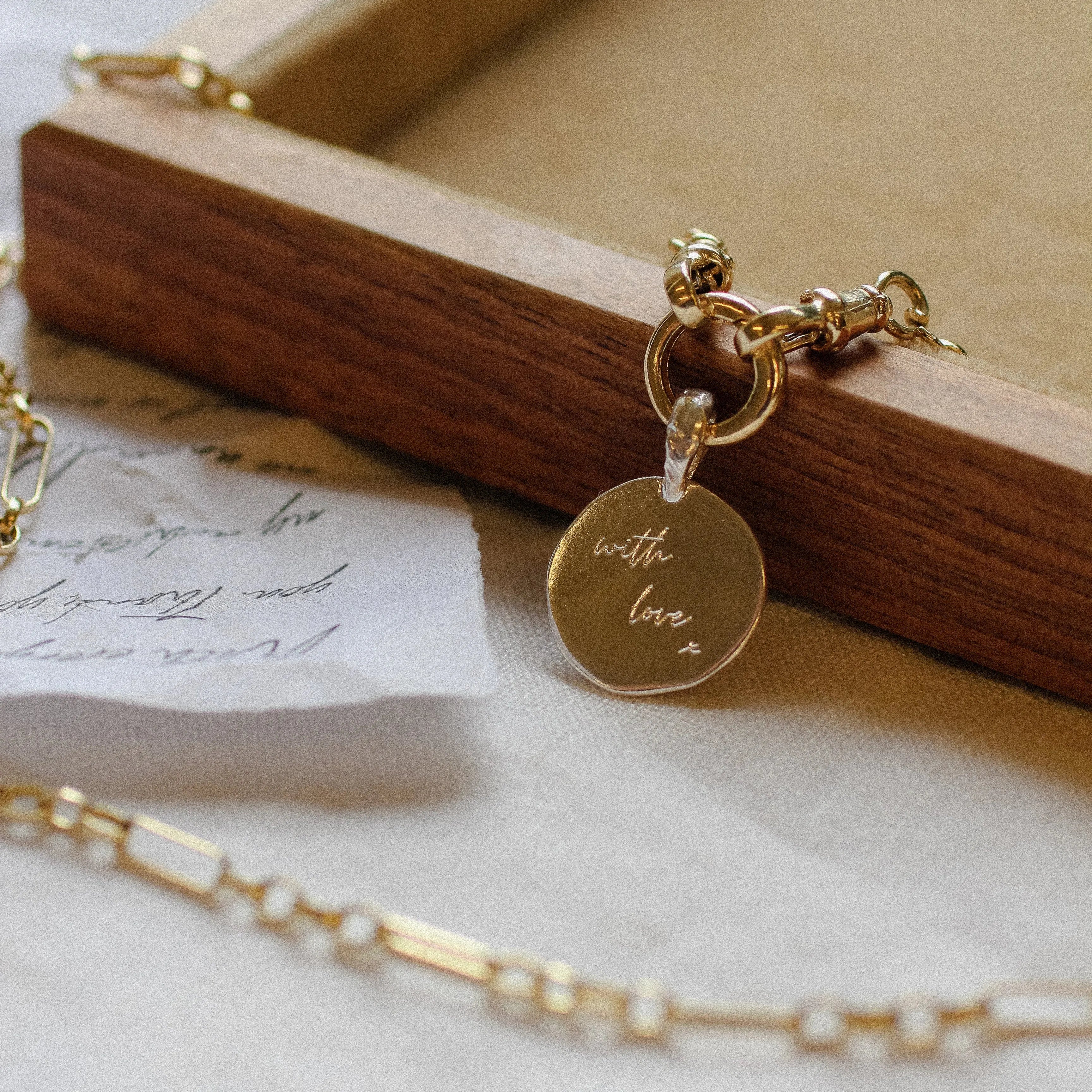Gold necklace pendant on a chunky chain with a 'with love' charm on a wooden box with a white background