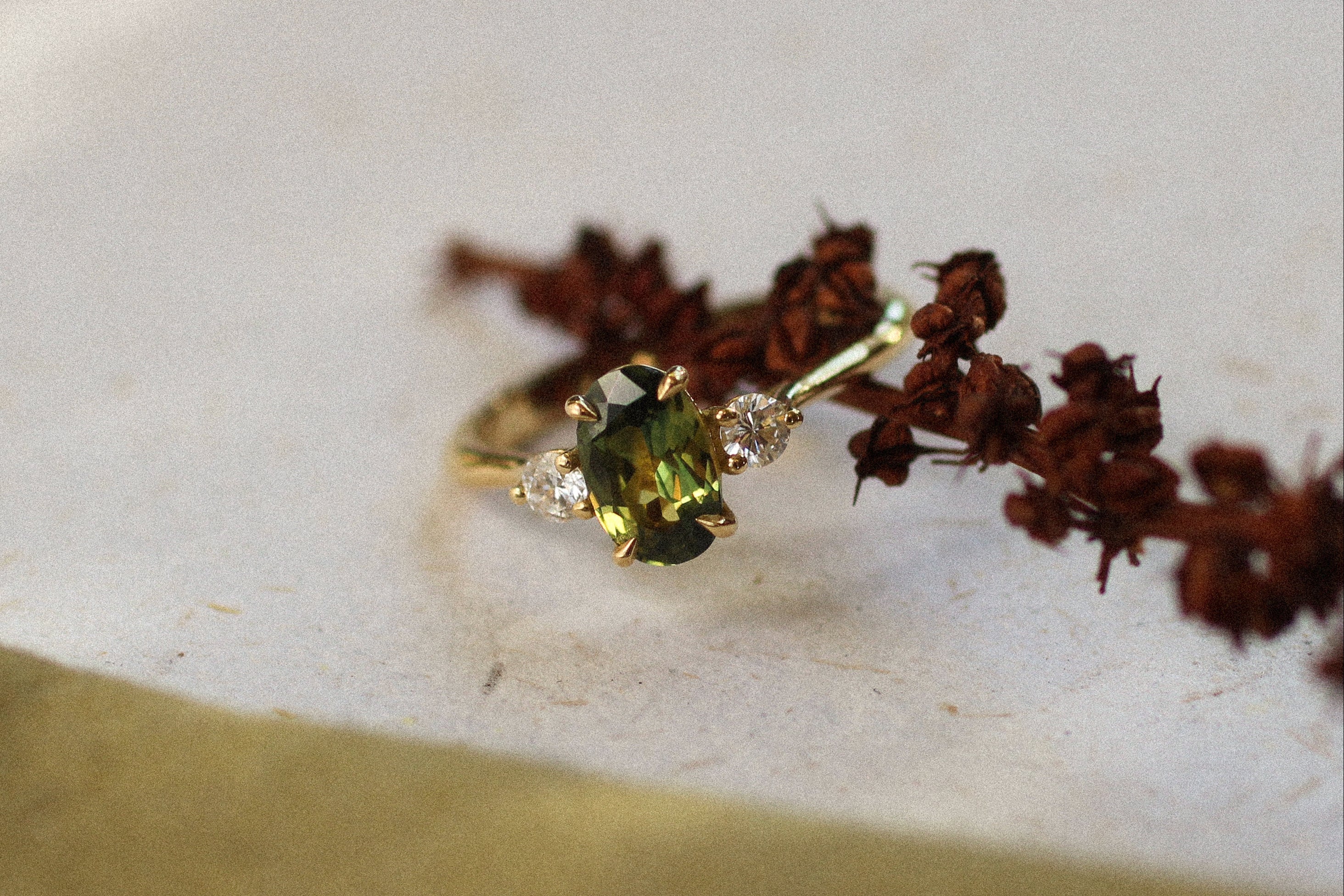 sapphire and diamond trilogy ring with a green gemstone on a white surface with dried flowers.