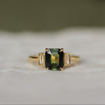 Gold ring with a green rectangular gemstone set with claws with 2 bezel set baguette diamonds on a white fabric background