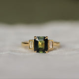 Gold ring with a green rectangular gemstone set with claws with 2 bezel set baguette diamonds on a white fabric background